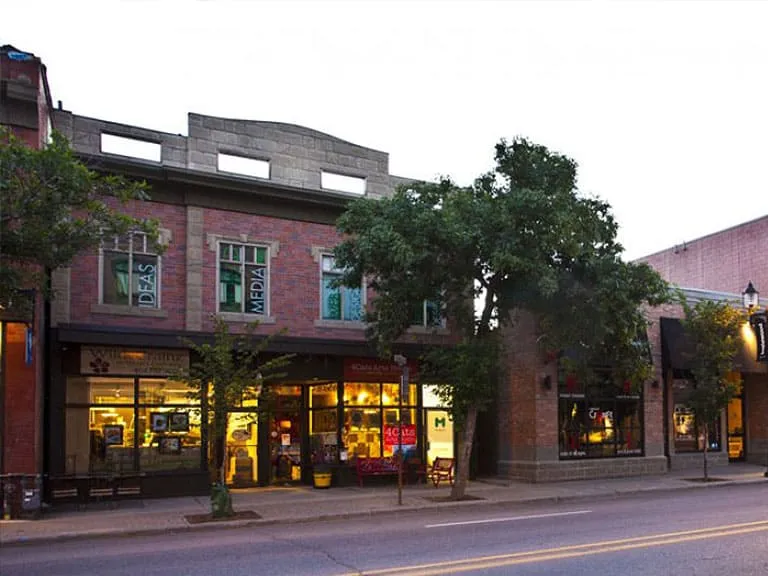 Shops in a brick building with warm lit windows and trees along the sidewalk